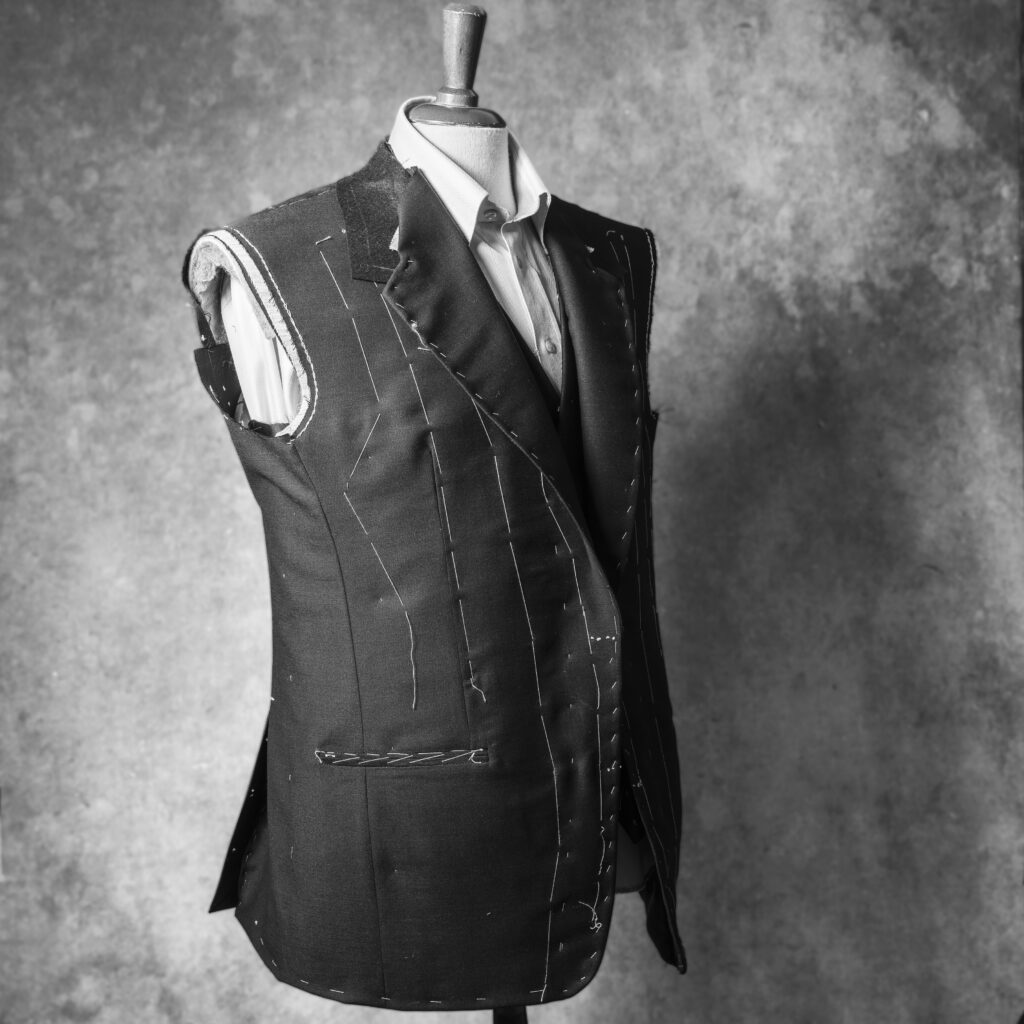 Mustavalkoisessa valokuvassa mallinuken torson päällä keskeneräinen miesten takki.